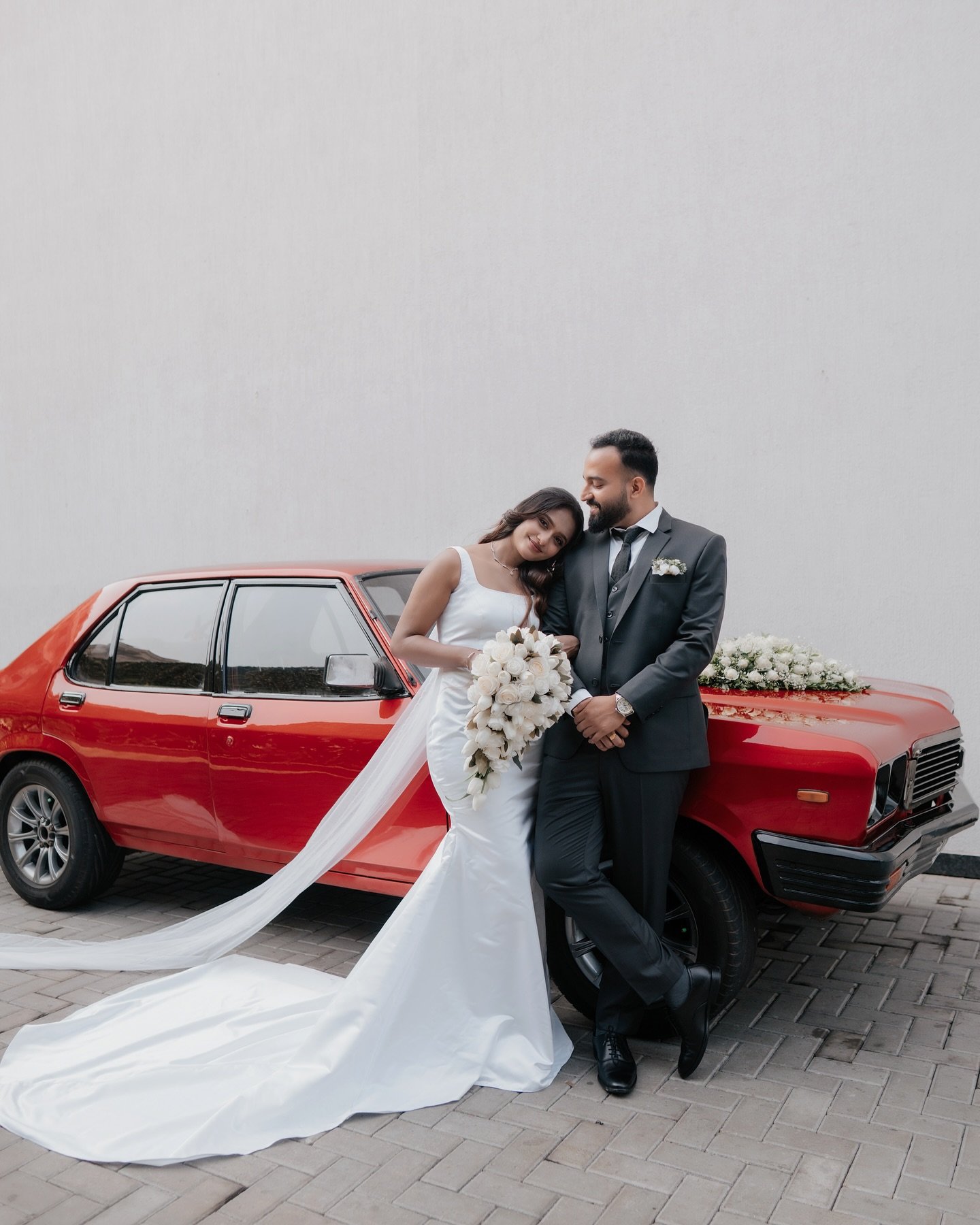 A newlywed couple posing together beside a red vintage car